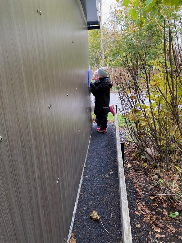 Två barn står vid en byggnad och de har buskar bakom sig.